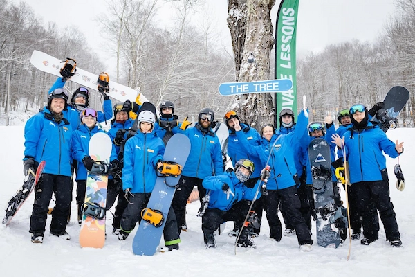 A large group of smiling Jiminy Peak® Mountain Resort employees, all wearing matching blue jackets, poses for a group photo in the snow while holding snowboards and skis.