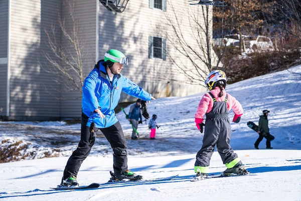 A ski instructor from KidsRule Mountain Camp is teaching a young child how to ski on a snowy slope. The instructor is leaning forward and pointing while the child listens.