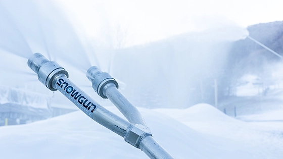 A close-up of a snow gun with two nozzles spraying snow on a mountain slope. The word "SNOWGUN" is visible on the equipment.