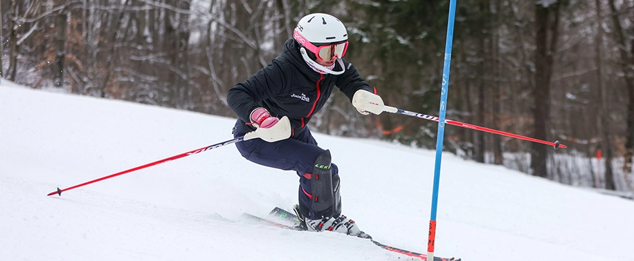 A focused young skier wearing a white helmet and goggles races down a snowy slope, carving a turn between slalom gates as part of the Tri-State program.
