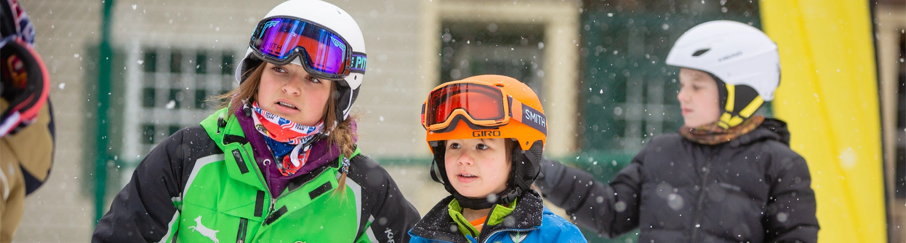 Three children in ski helmets and goggles are looking forward attentively during a lesson, with snow falling around them.