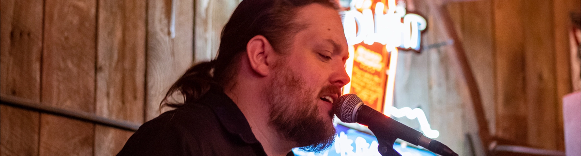 A close-up of a bearded man with a ponytail singing into a microphone under warm, dim light, set as the website banner for the Events Calendar.