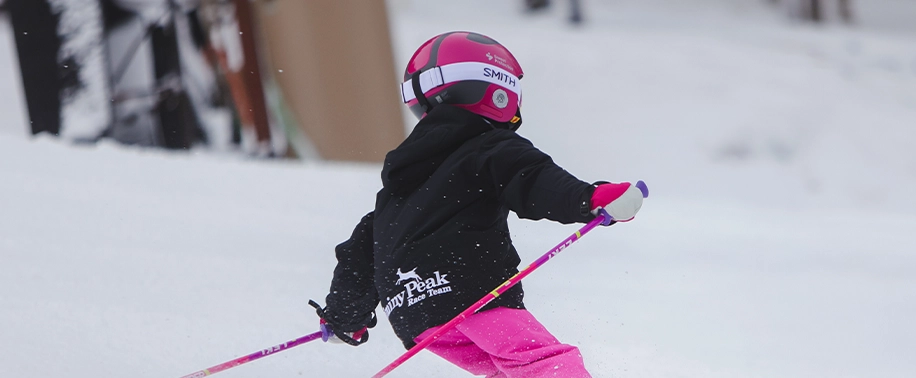 A young skier in bright pink pants, a black Jiminy Peak Race Team sweatshirt, and a pink helmet practices skiing down a snowy slope. The image is a call-to-action for the Alpine Racing Development Program.